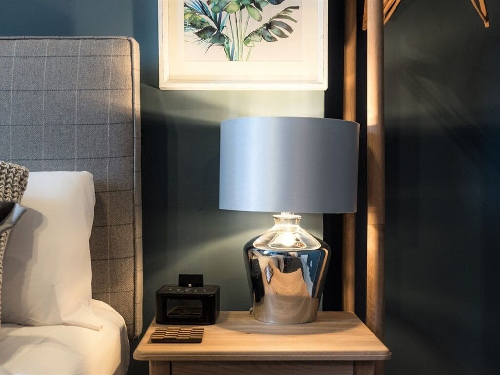 A modern lamp on a bedside table next to a bed with a grey headboard.