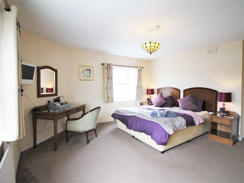 A bedroom with a bed, desk, chair, bedside table, lamp, and a window.