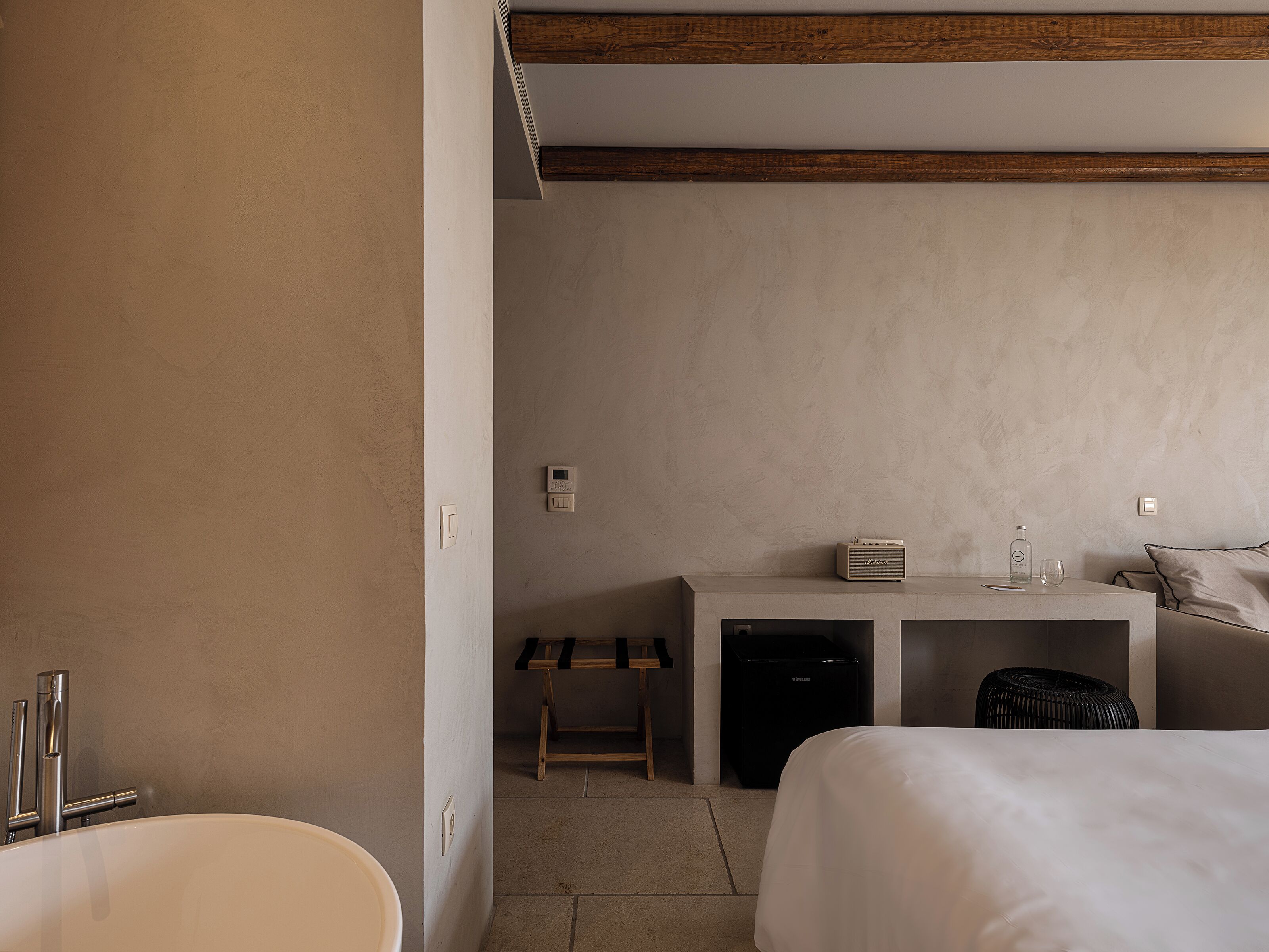 A hotel room with a bed, a desk with a computer, a small stool, a black trash bin, and a white sink.