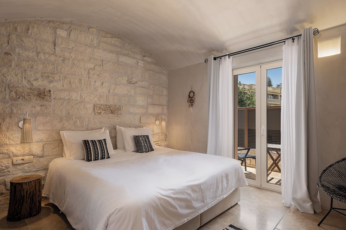 Un dormitorio con pared de piedra, una cama con ropa de cama blanca, un taburete de madera pequeño y vistas a un balcón exterior.