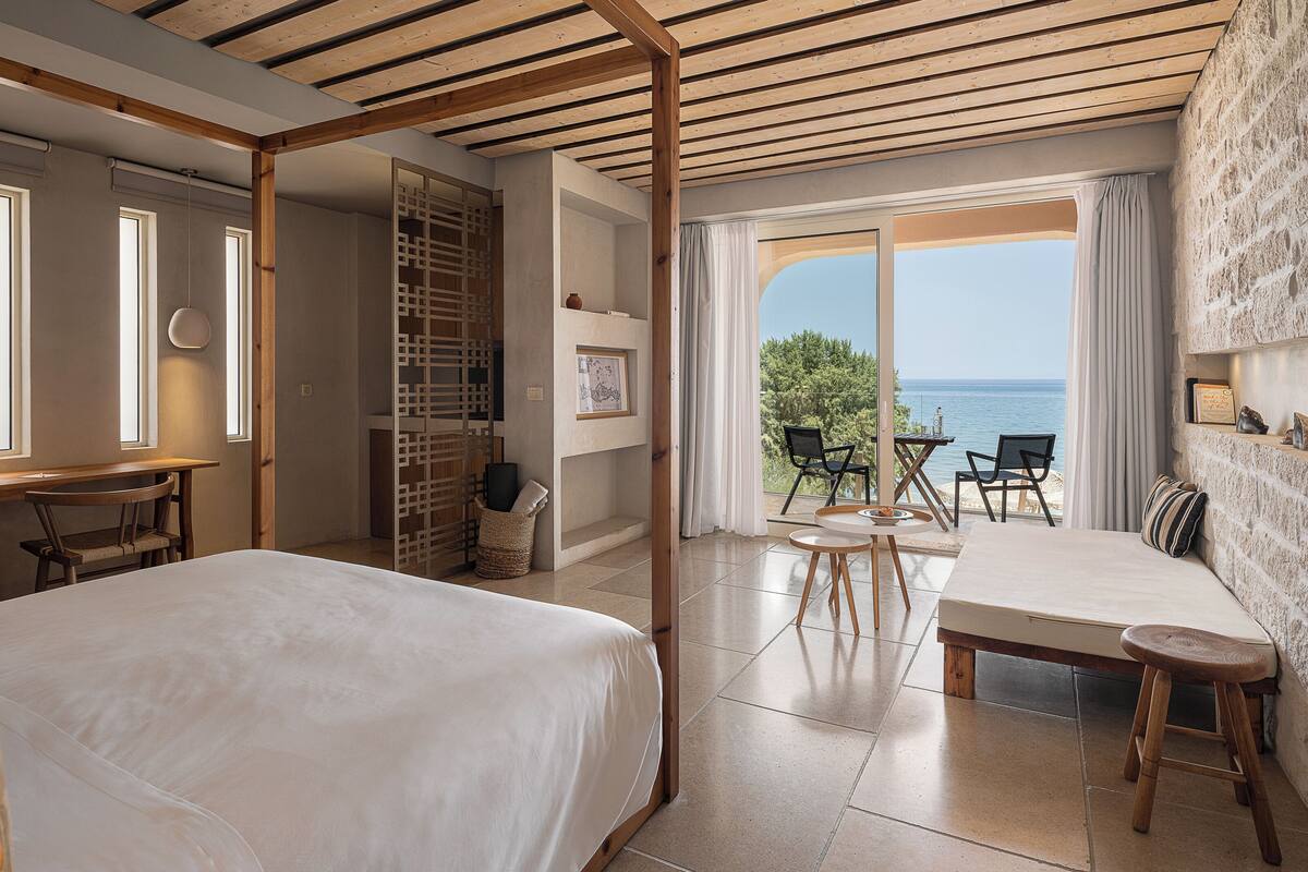 Un dormitorio con una cama grande, una mesa de comedor y sillas. Se ve el mar a través de las puertas del balcón.