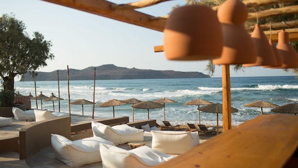 Um espaço de relaxamento à beira-mar, com assentos acolchoados em branco e guarda-sóis de palha, com vista para o mar.