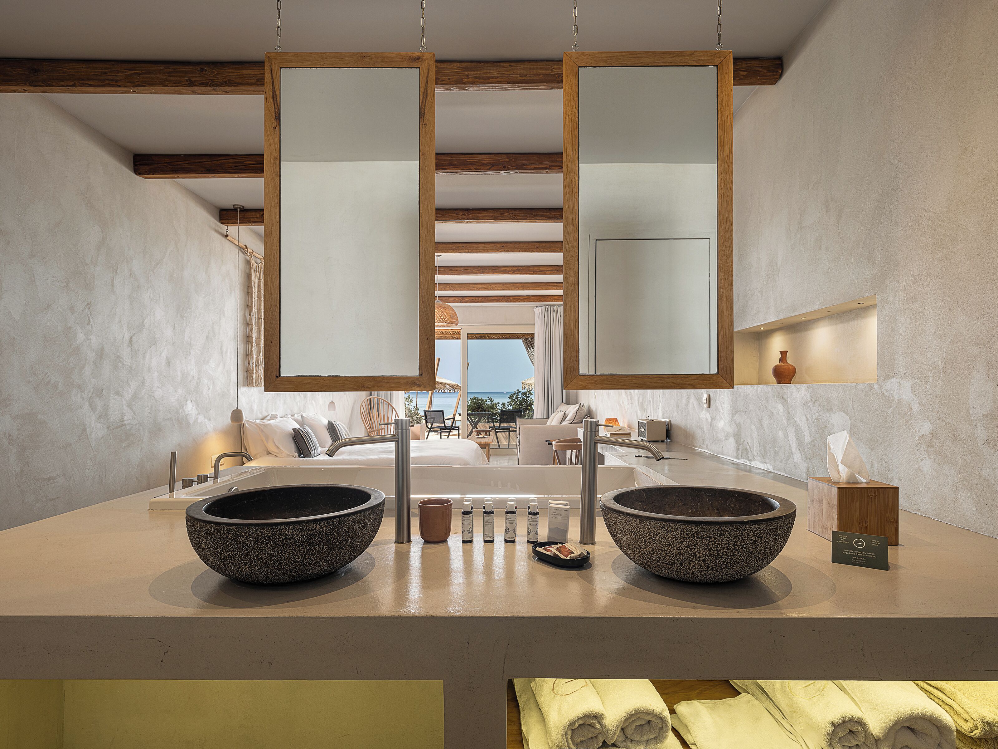 A modern bathroom with two large stone basins, a wooden ceiling, and a view of the sea.