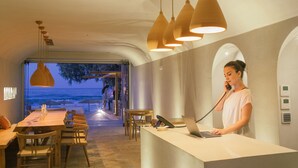 Bancada de recepção com um laptop, um telefone e uma mesa de madeira com cadeiras. Há luminárias pendentes sobre a mesa.