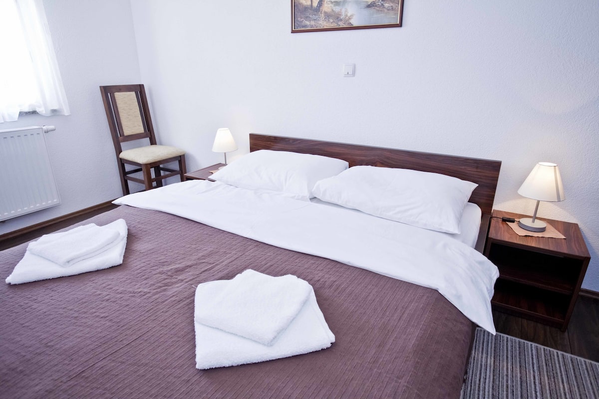 Un lit bien fait, avec du linge de lit blanc, une table de chevet en bois sur laquelle se trouve une lampe, une chaise et un tableau accroché au mur.