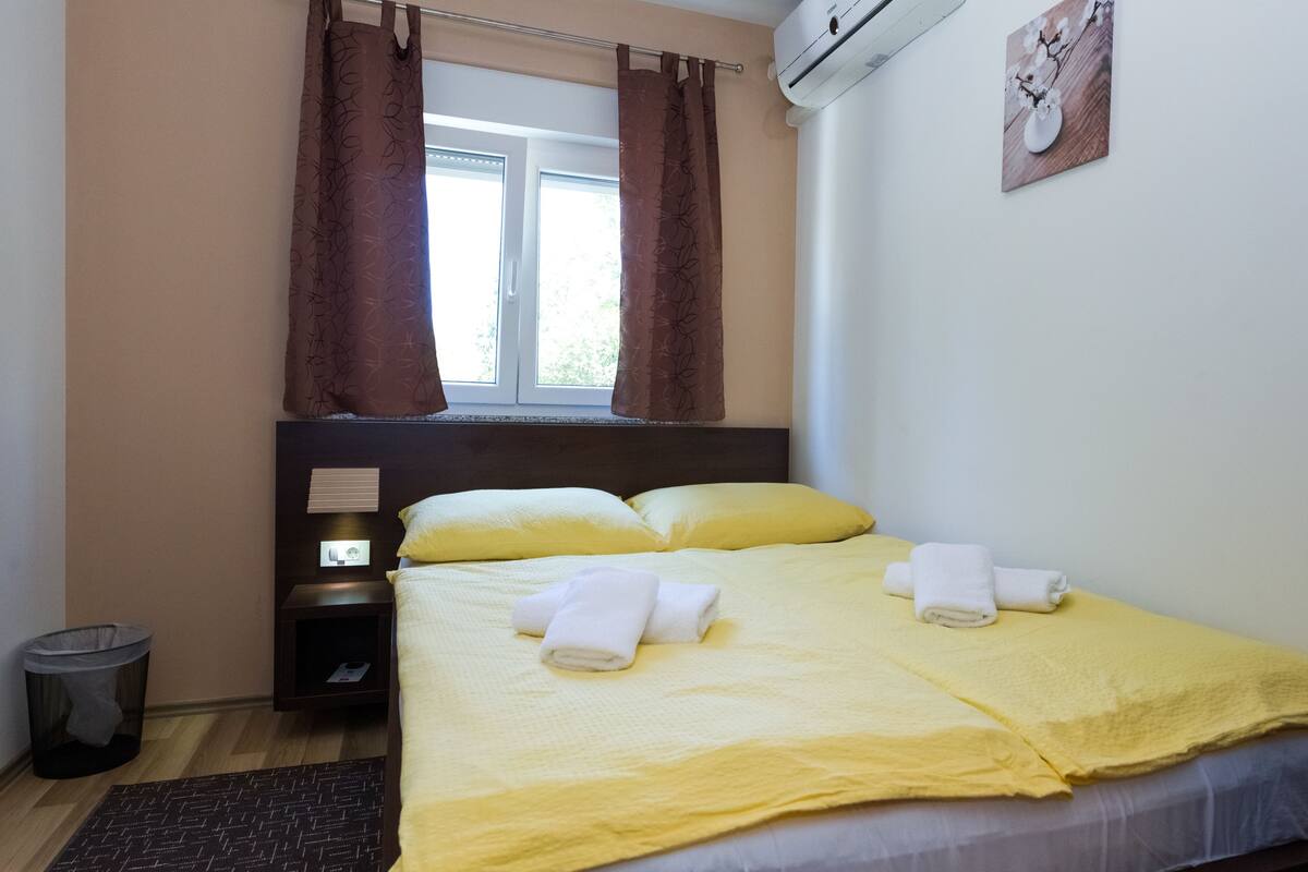 Une chambre d’hôtel avec un lit, deux oreillers, une table de chevet, une poubelle et une fenêtre avec des rideaux.