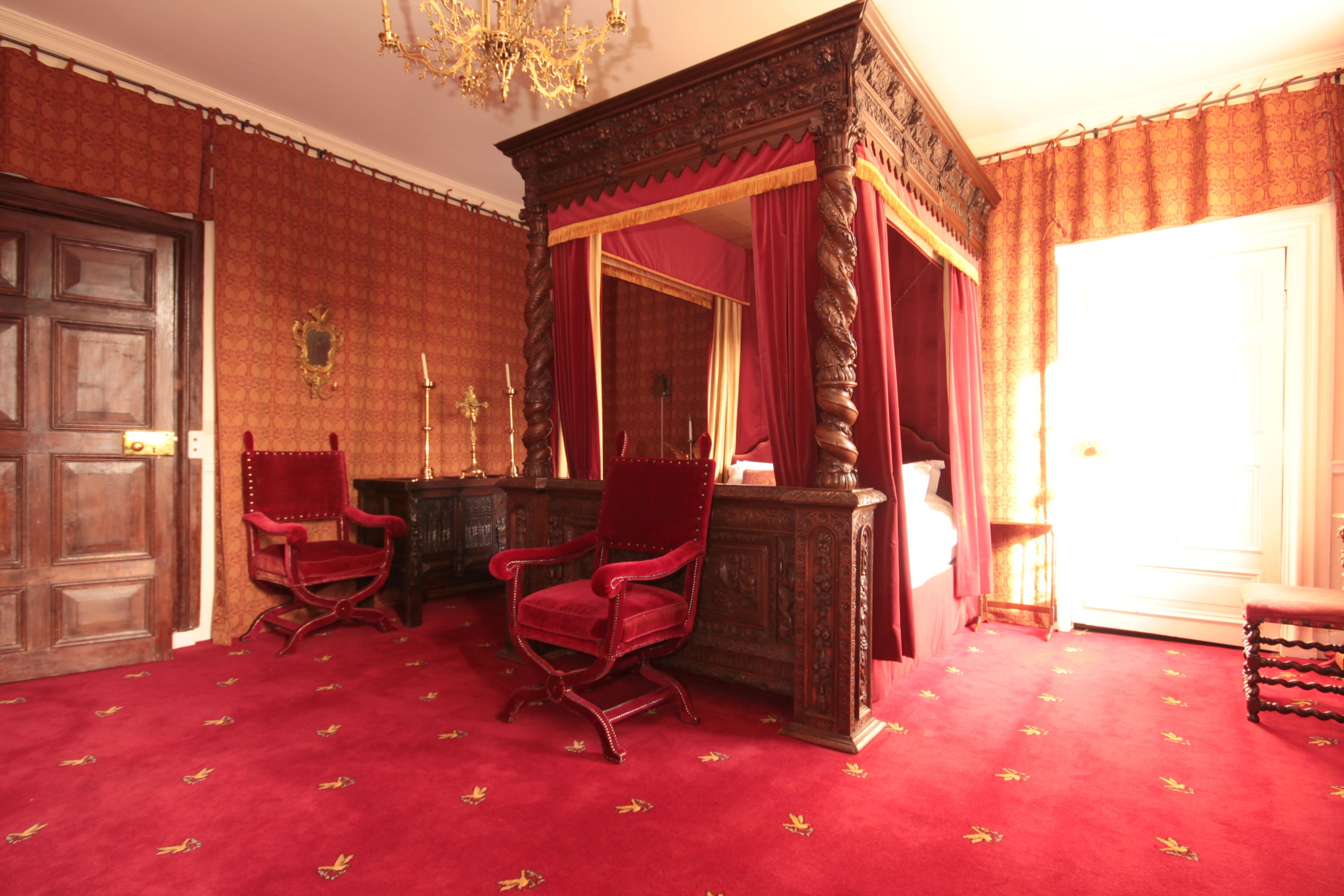 Une chambre dotée d’un lit à baldaquin, de deux fauteuils à revêtement rouge, d’un bureau en bois et d’un grand miroir.