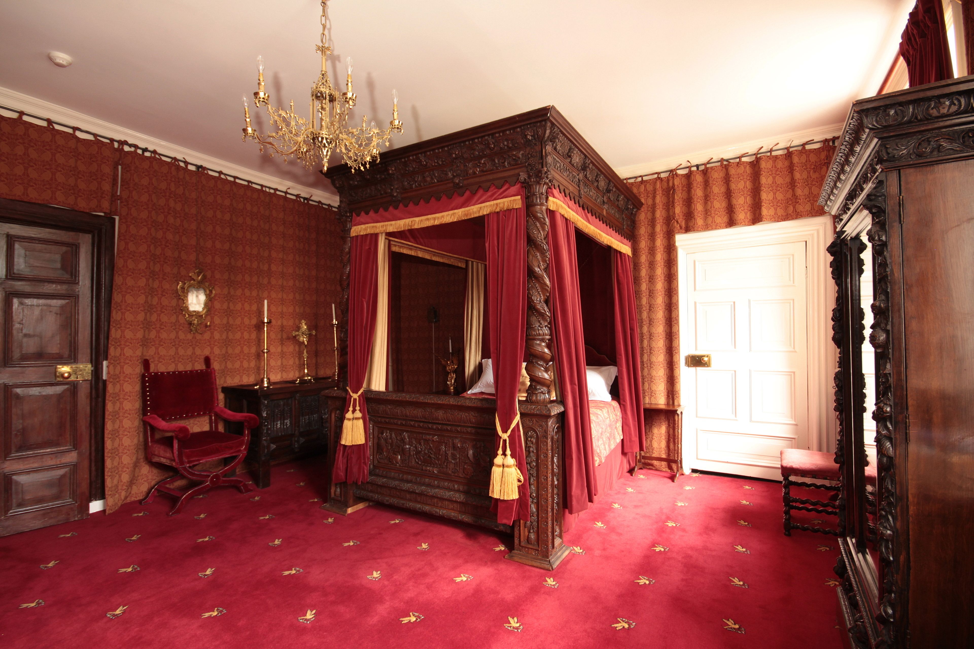 A room with a four-poster bed, a chandelier, a red carpet, and a wooden door.
