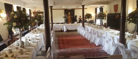 Un salón de banquetes con largas mesas dispuestas para un evento formal, que cuenta con un pasillo central y un techo ornamentado.