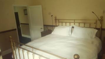 Una cama con estructura metálica y ropa de cama blanca en una habitación con puerta y una mesita de noche pequeña.