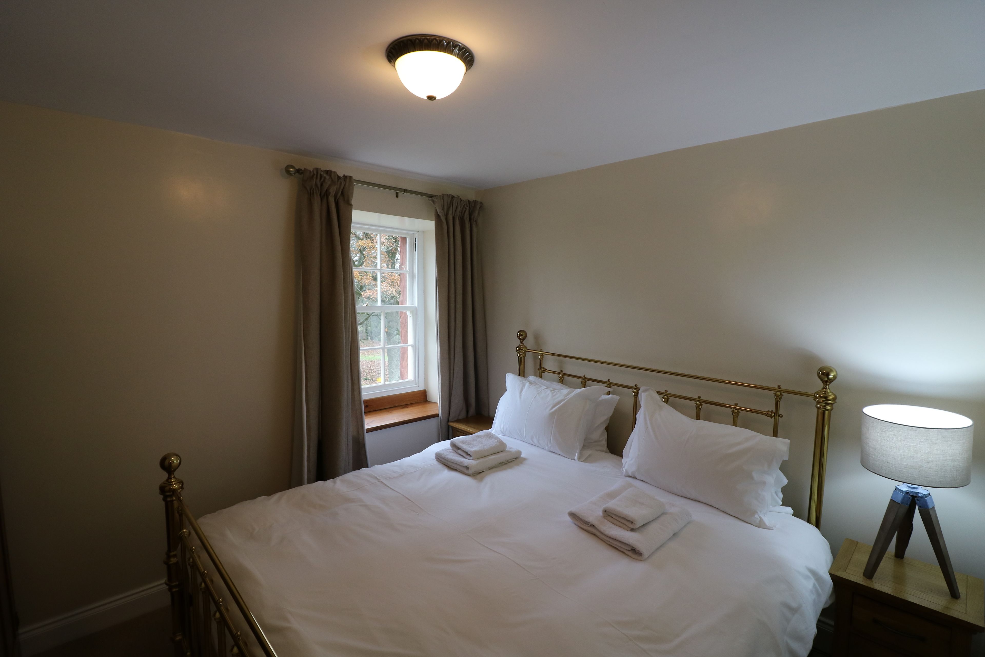 A neatly made bed with white linens and pillows, a bedside lamp, and a window with curtains.