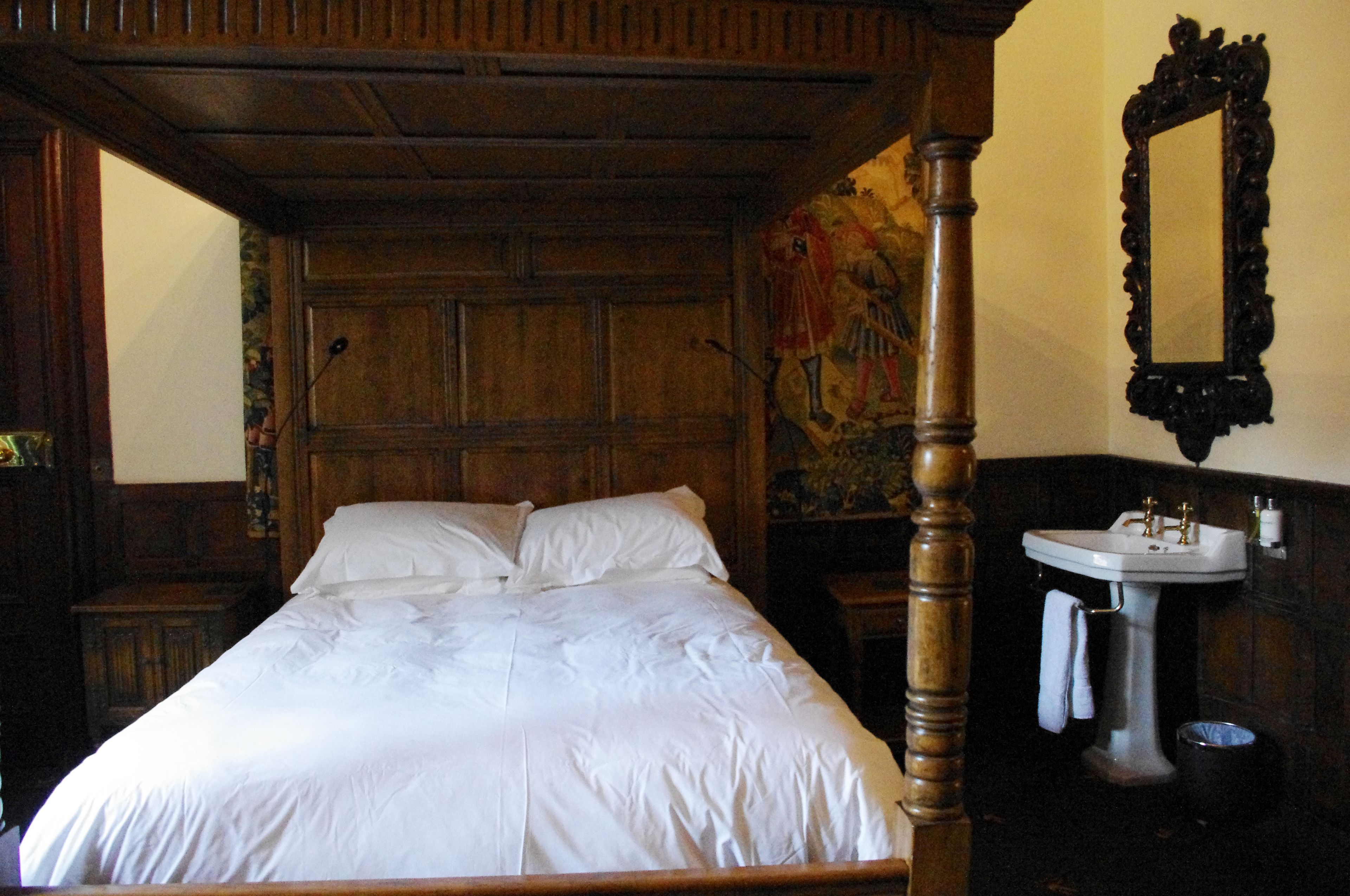 A traditional wooden four-poster bed with white linens, a wooden nightstand, and a wall-mounted mirror.