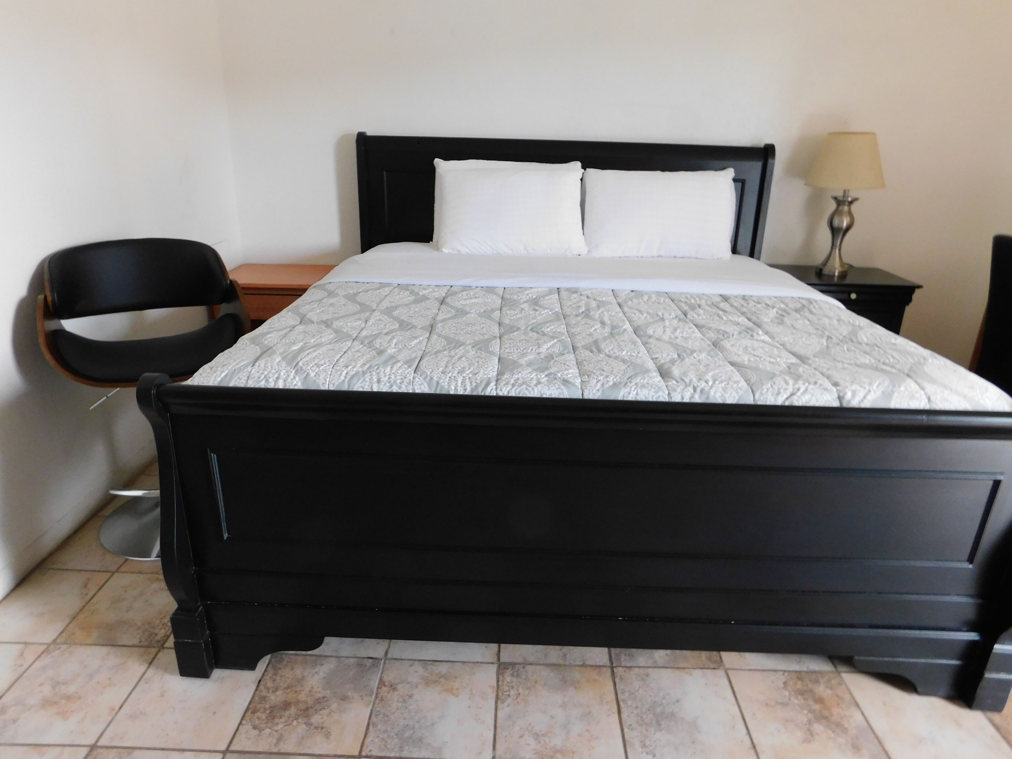A bed with a black frame, a white mattress, and a patterned bedspread. There is a chair and a nightstand with a lamp.