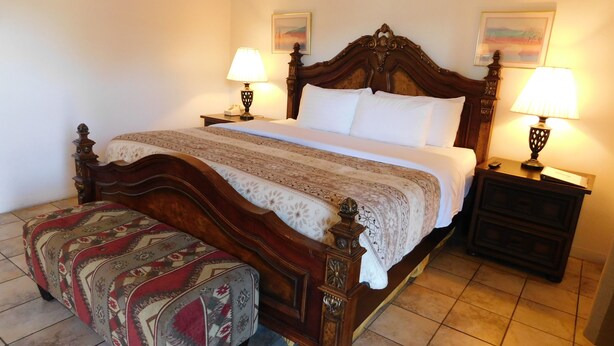 Una cama bien hecha con cabecera de madera, dos mesitas de noche con lámparas, un otoman estampado y dos cuadros enmarcados en la pared.