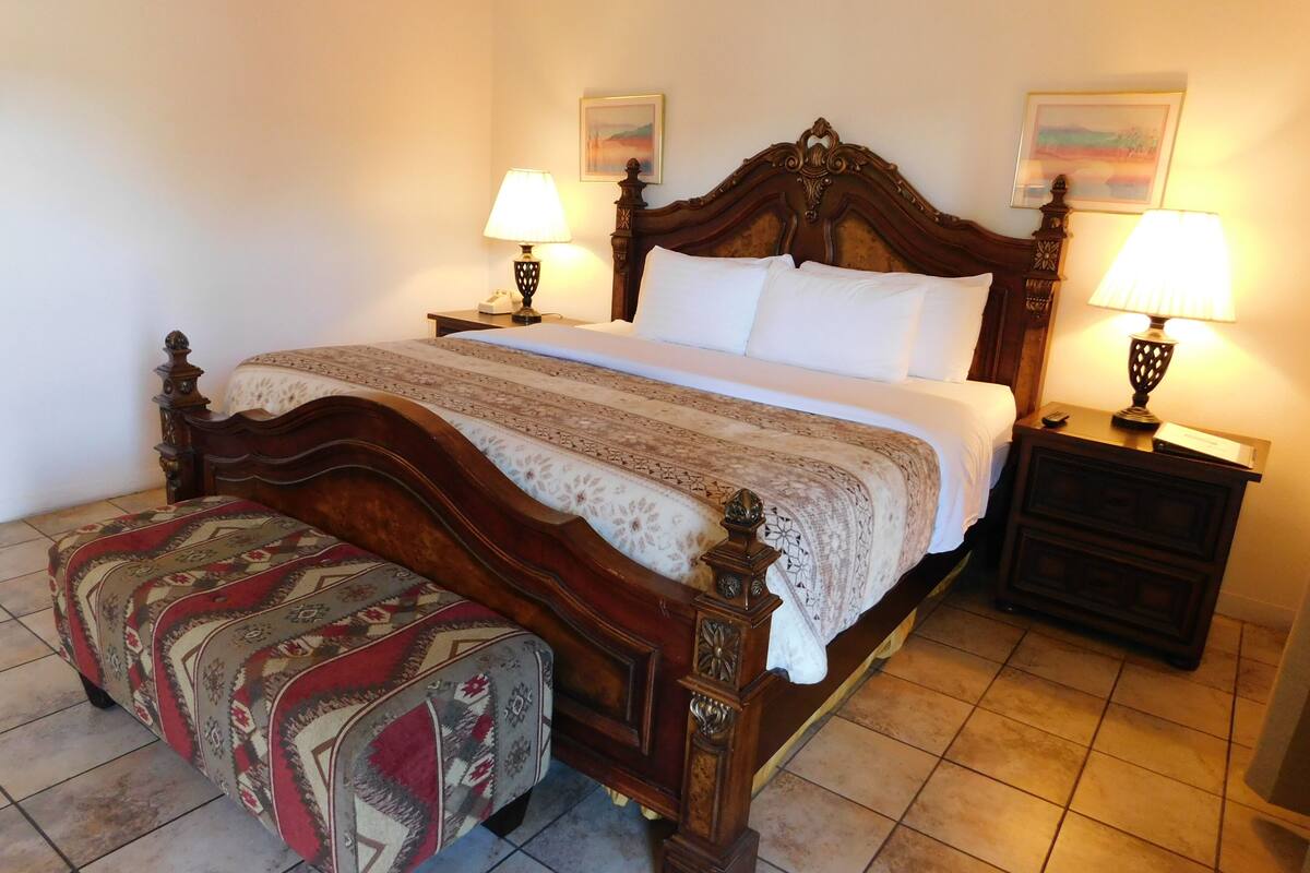 A neatly made bed with a wooden headboard, two bedside tables with lamps, a patterned ottoman, and two framed pictures on the wall.