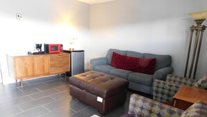 Una sala de estar con sofá, reposapiés, sillón estampado, y un mueble de madera con microondas y cafetera.
