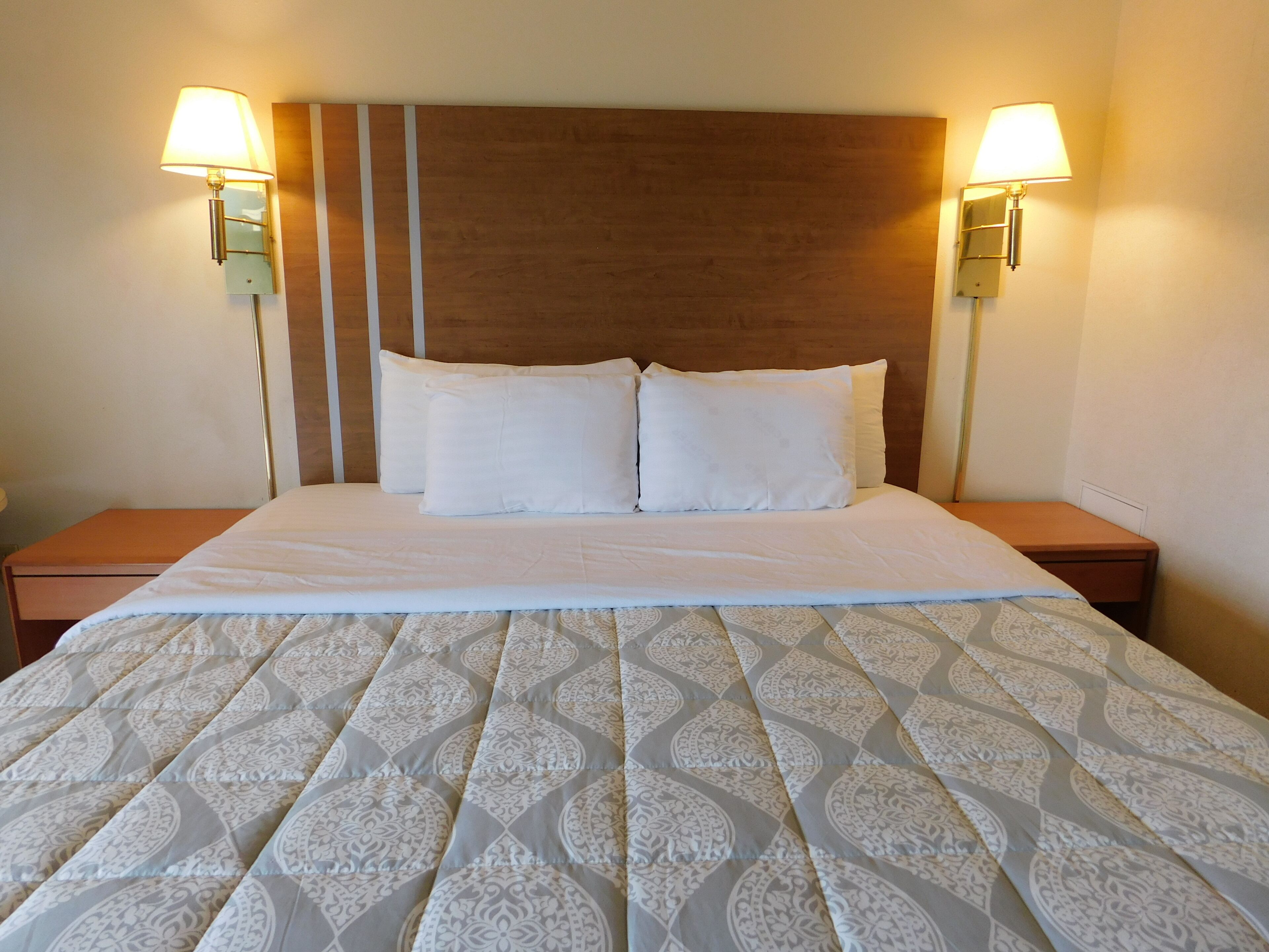 Un lit bien fait, recouvert d’une couvre-lit à motifs, avec deux oreillers blancs et une tête de lit en bois. Deux lampes de chevet sont placées de chaque côté du lit.