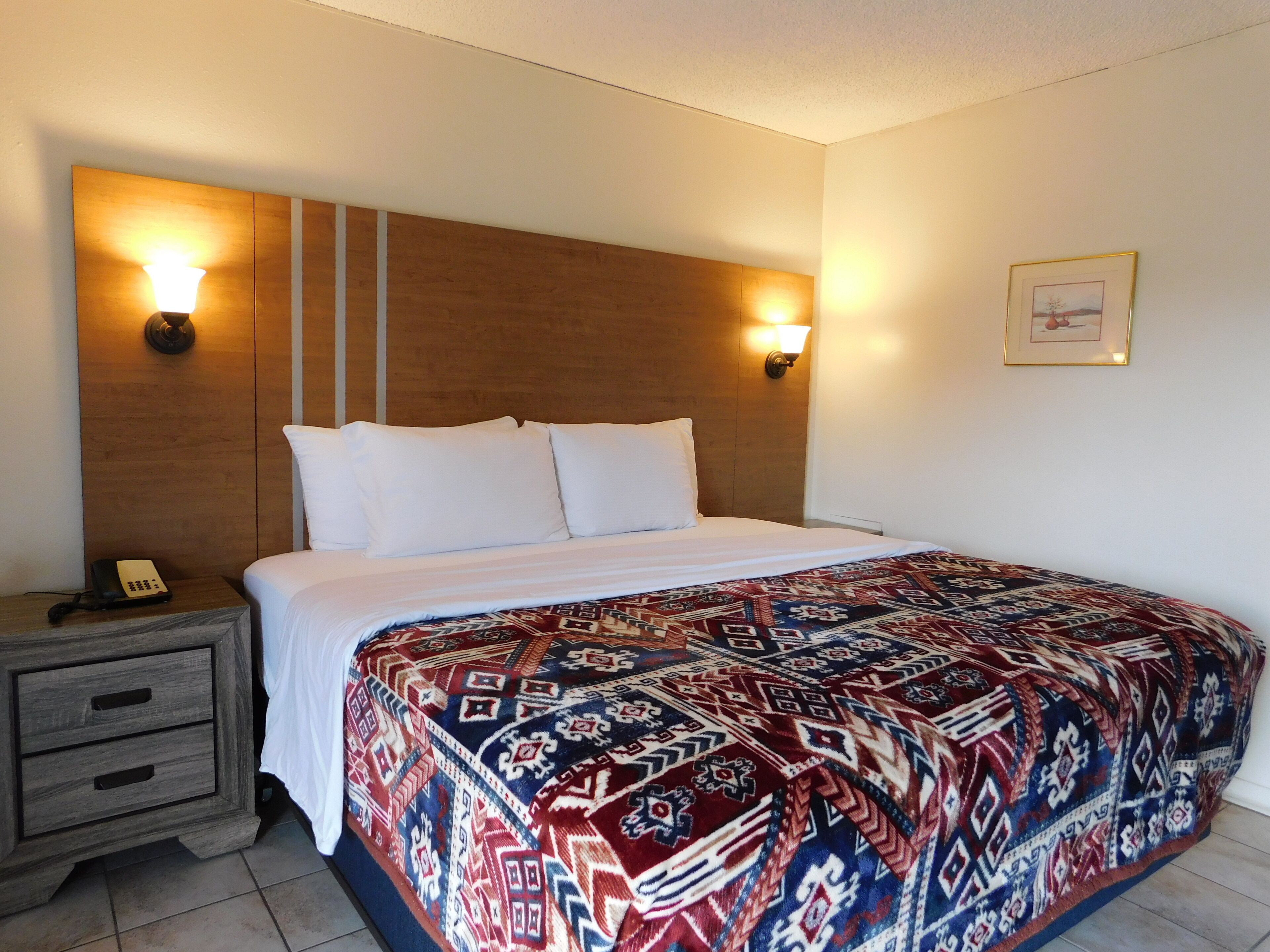 A hotel room with a large bed, two bedside lamps, a nightstand with a phone, and a framed picture on the wall.