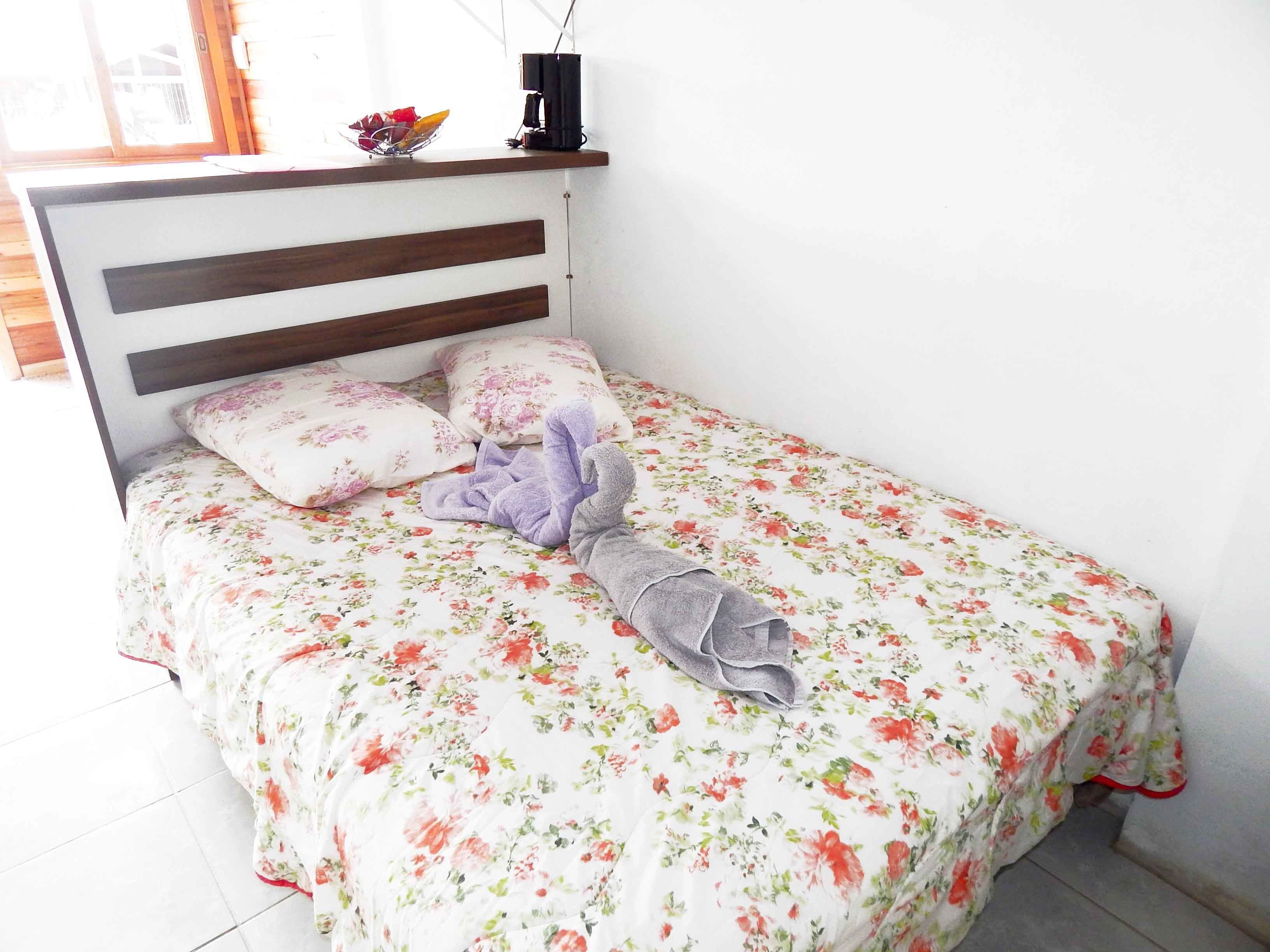 A neatly made bed with floral bedding, a wooden headboard, and a bedside table with a coffee maker and a bowl of fruit.