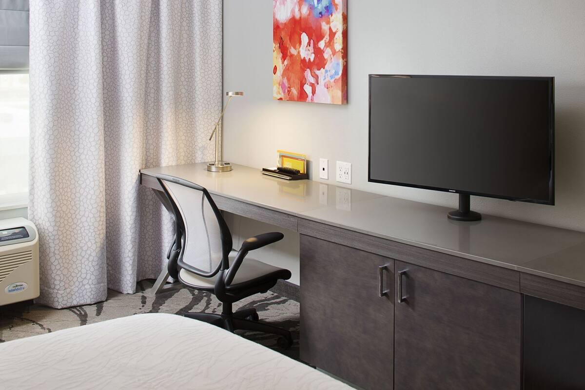A hotel room with a desk, chair, TV, and a bed.