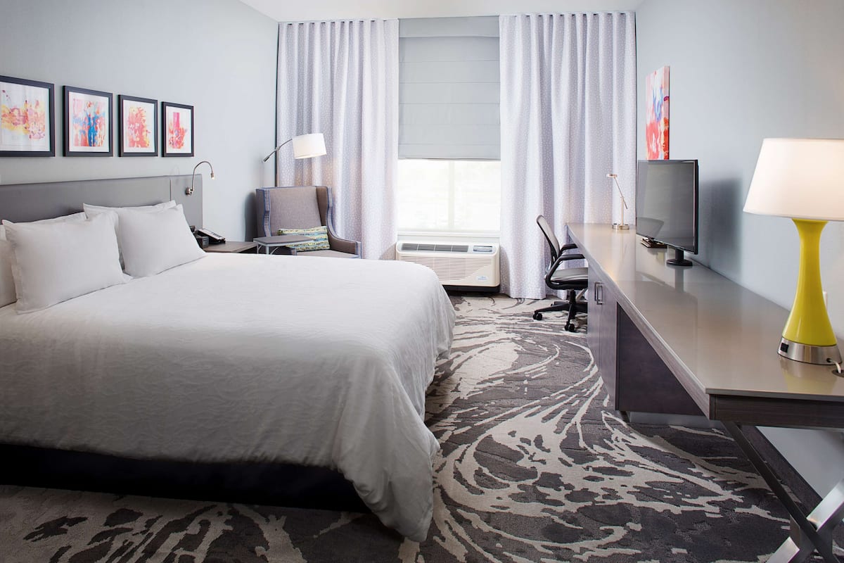 A hotel room with a large bed, a desk with a TV, a chair, and a desk lamp. There are curtains on the windows and a rug on the floor.
