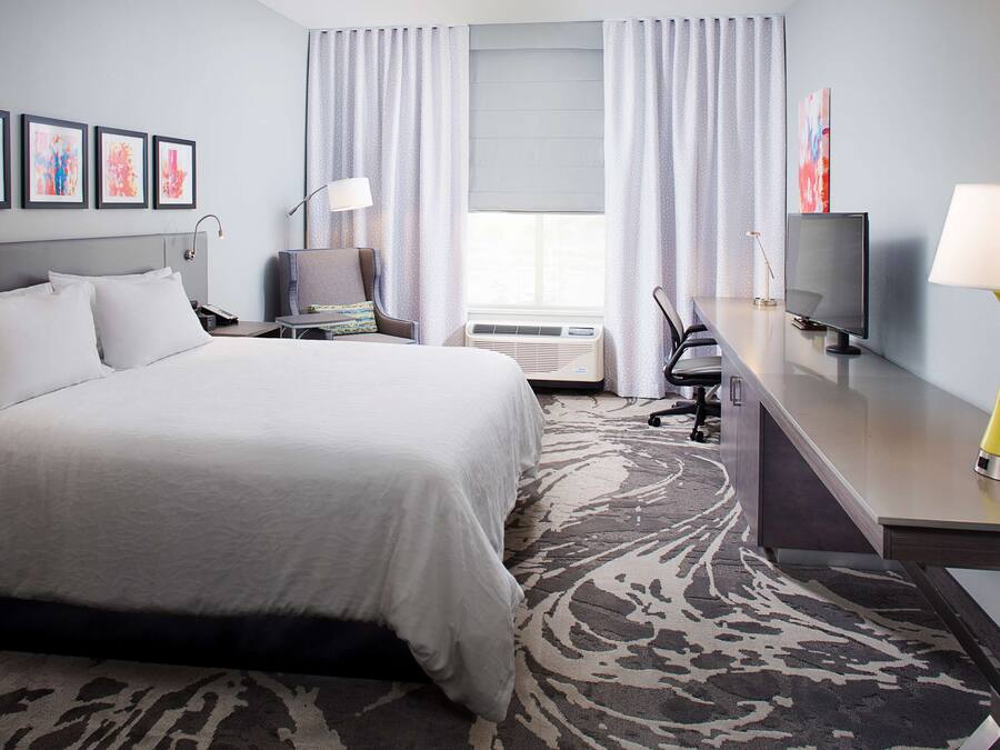 A hotel room with a large bed, a desk with a TV, a chair, and a desk lamp. There are curtains on the windows and a rug on the floor.