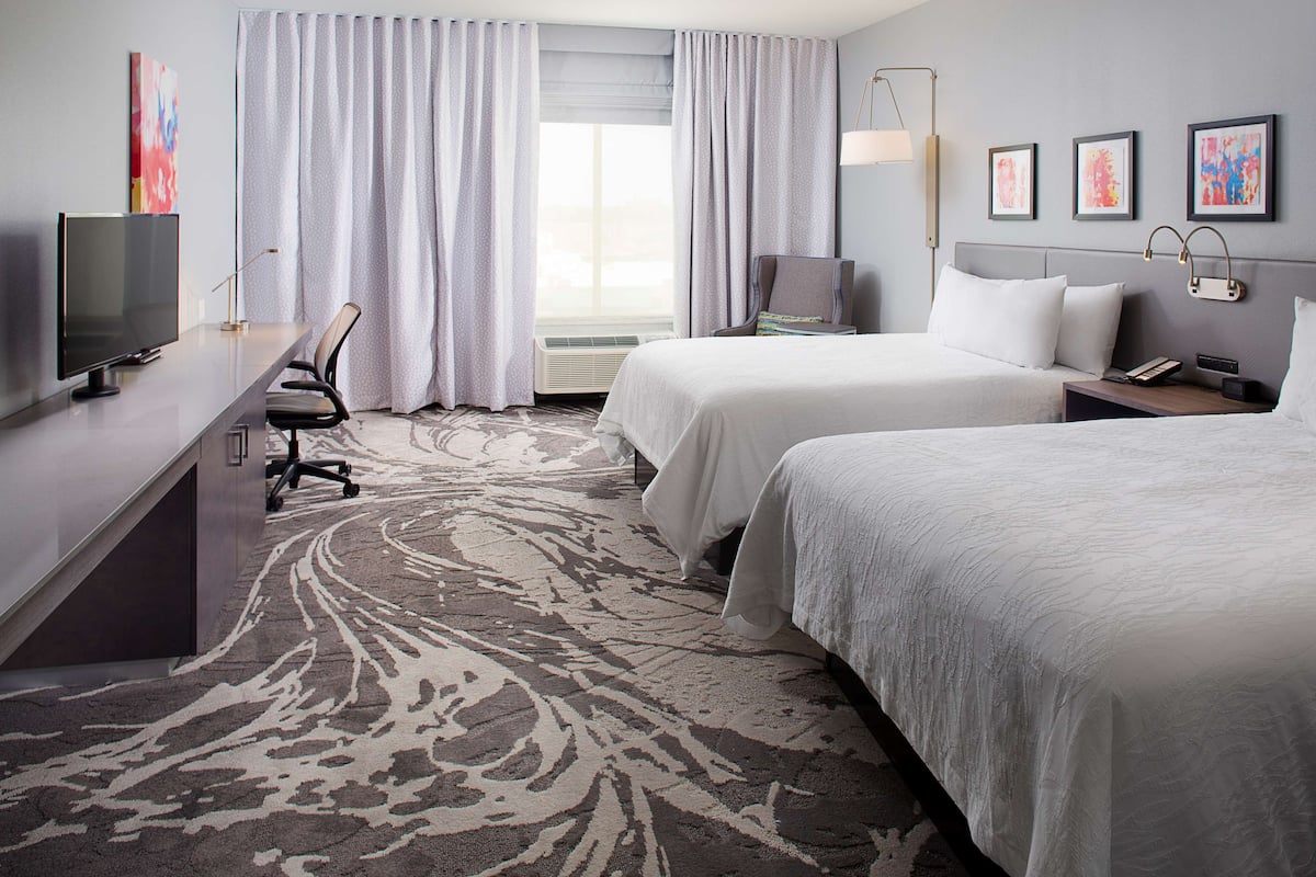 A hotel room with two beds, a desk with a TV, a chair, and a large window with curtains.