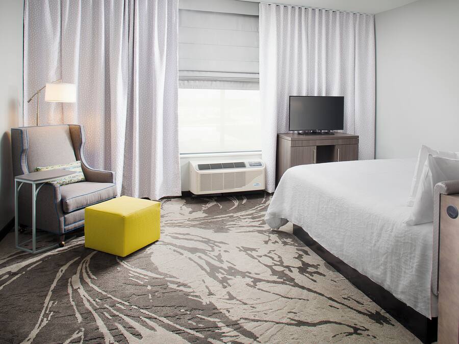 Une chambre d’hôtel avec un lit, une télévision, une chaise, une petite table et une fenêtre avec des rideaux.