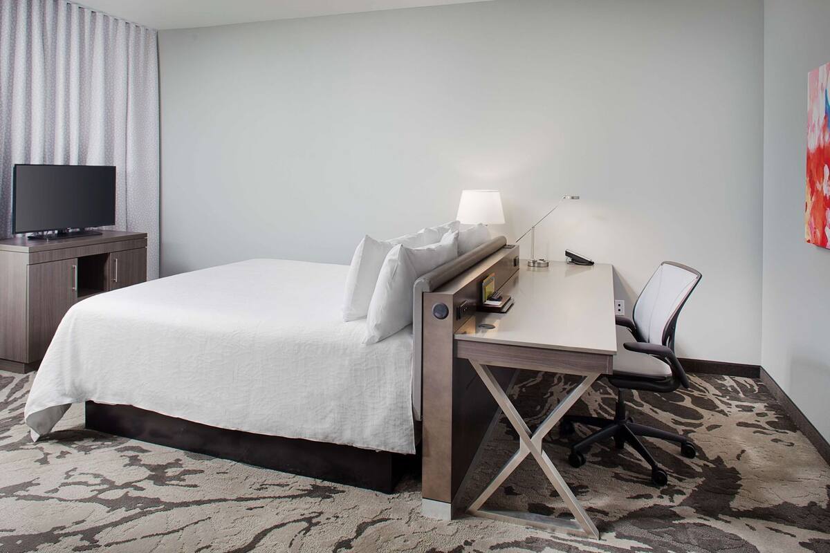 A hotel room with a bed, a desk with a lamp, a chair, a television, and a carpeted floor.