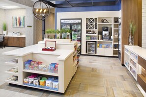 A modern coffee shop interior with a counter, shelves stocked with snacks, a coffee machine, and a refrigerated display of cold beverages. - Hilton Garden Inn Dallas/Arlington South (Arlington)