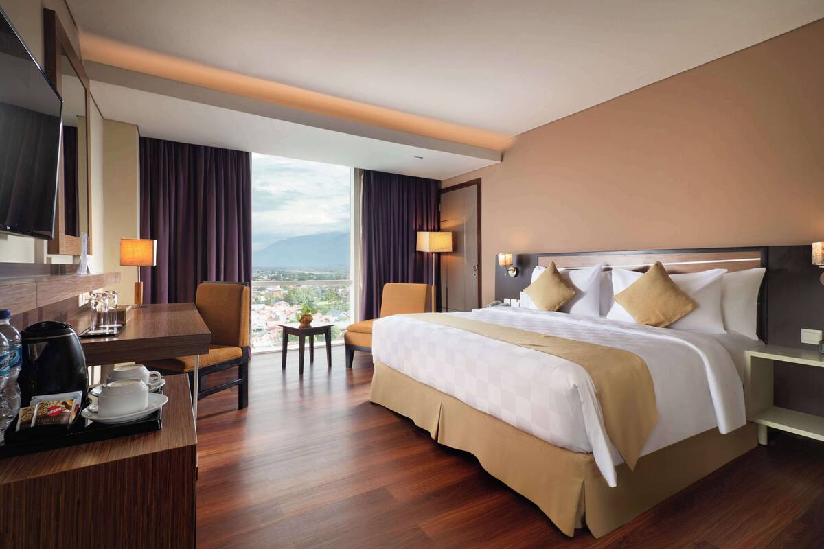 A hotel room with a large bed, a desk with a coffee maker, a chair, and a view of the city.