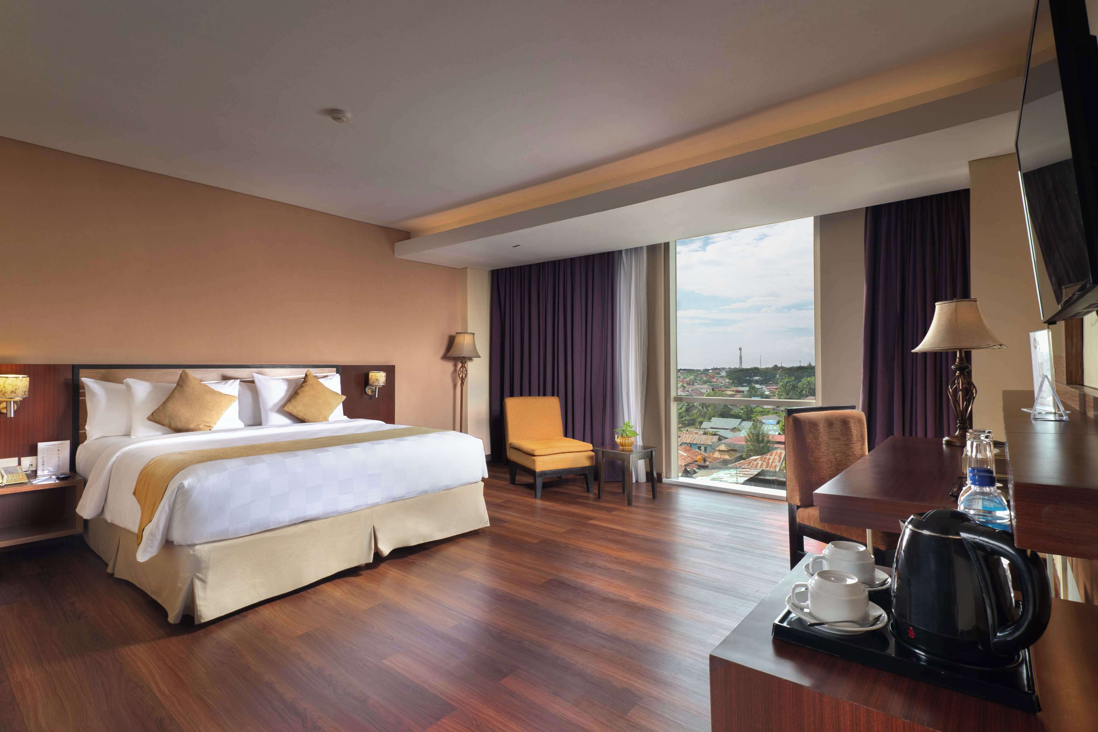 A hotel room with a large bed, a desk with a kettle, a chair, and a view of a cityscape through the window.