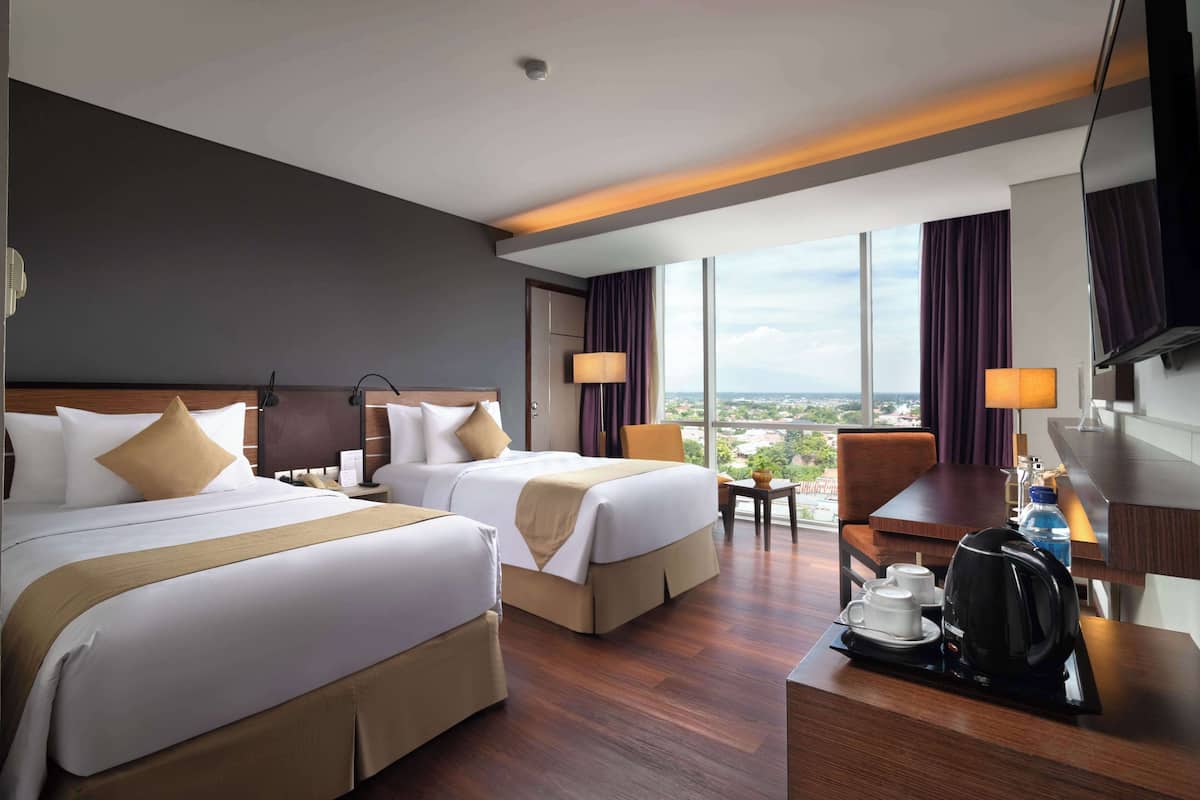 A hotel room with two beds, a desk with a coffee maker, a chair, and a large window offering a city view.