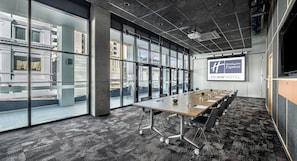 A conference room with a long table, chairs, a projector screen, and large windows offering a city view.