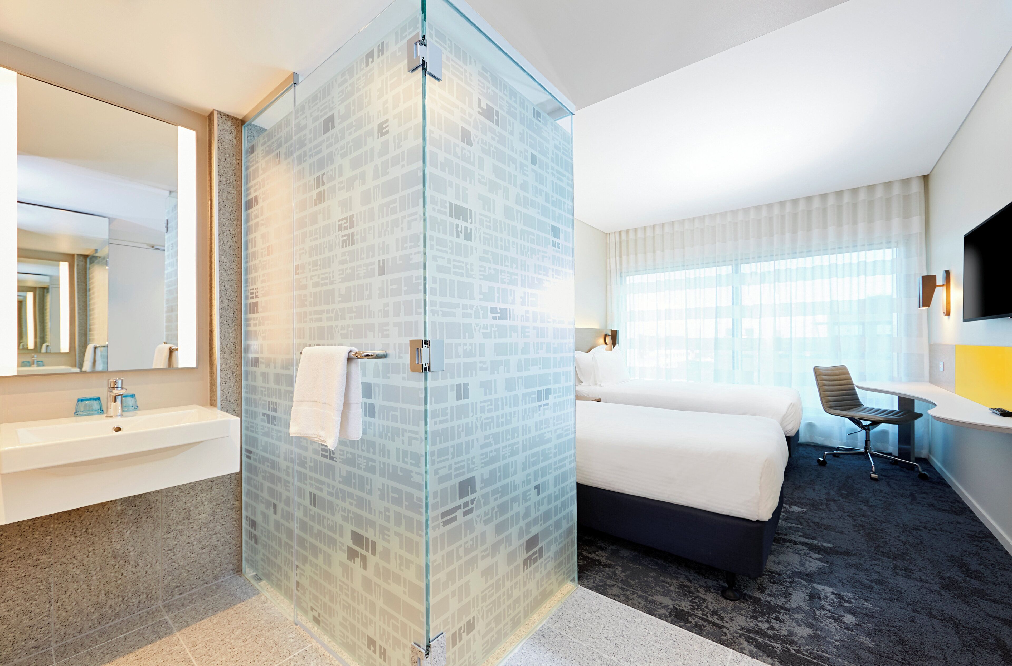 Habitación de hotel con cabina de ducha de vidrio, cama, escritorio, silla y televisor.