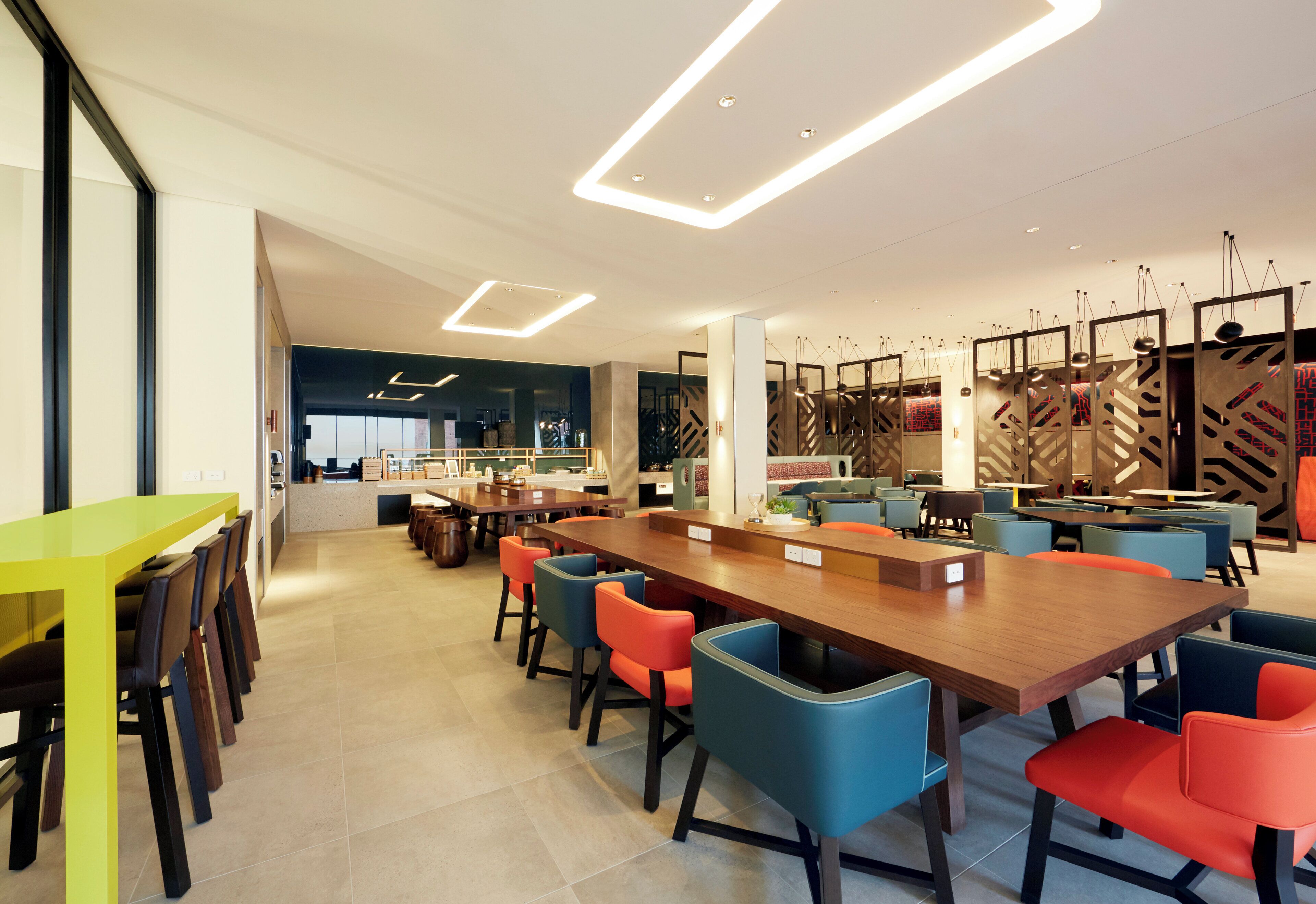 Un comedor moderno con una larga mesa de madera, sillas coloridas y una cocina al fondo.
