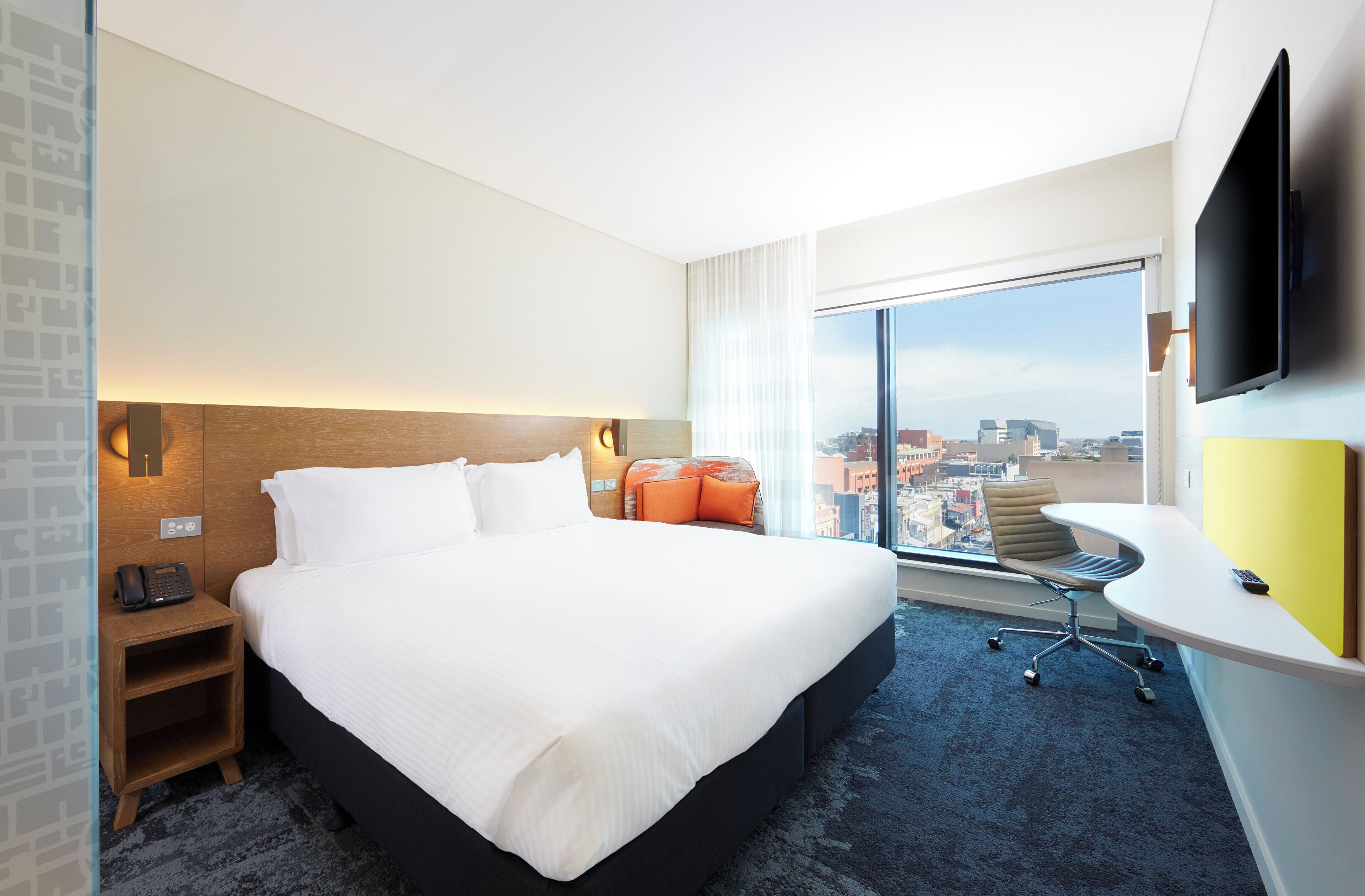 A hotel room with a large bed, a desk with a chair, a flat-screen TV, and a city view.