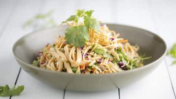 En skål med nudelsallad, olika grönsaker och koriander, placerad på en vit träyta.