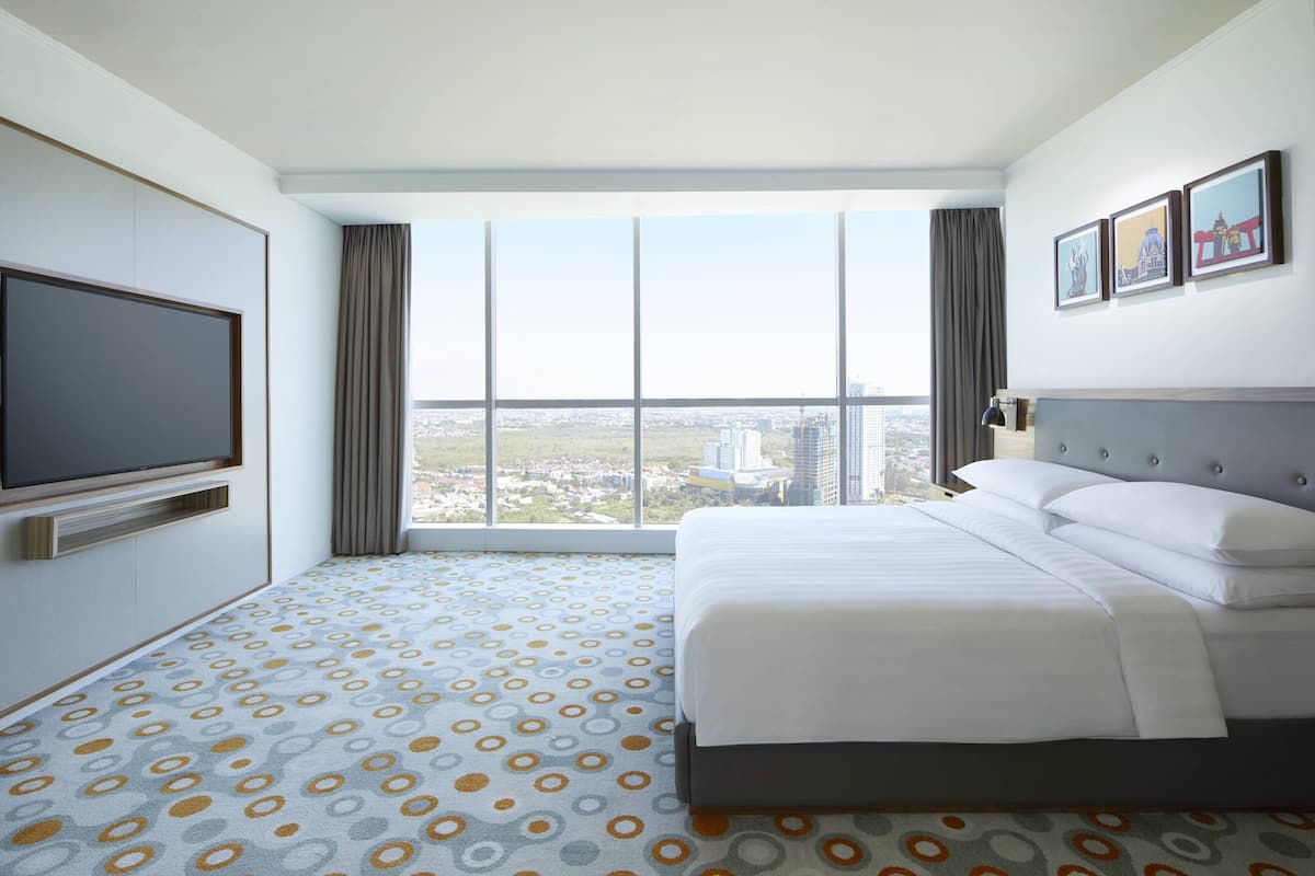 Habitación de hotel con una cama grande, un televisor de pantalla plana y vistas a la ciudad a través de amplias ventanas.