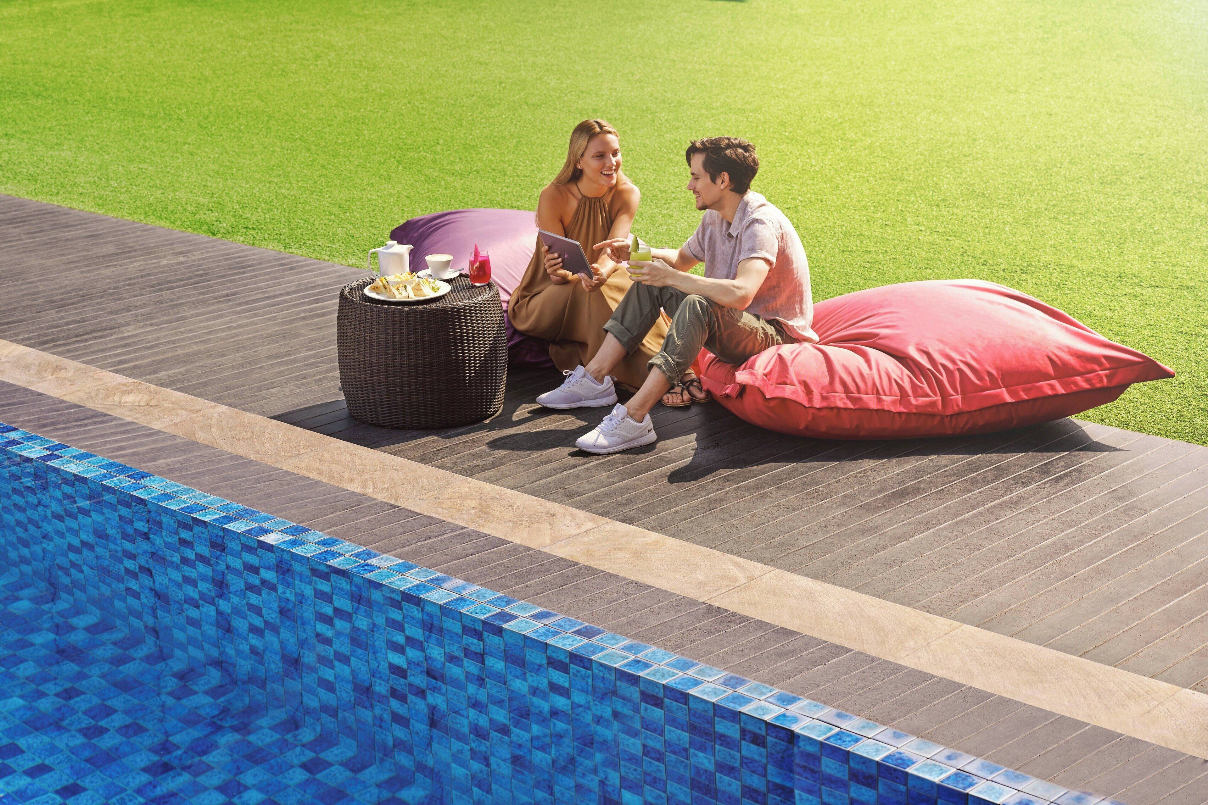 Dos personas sentadas junto a una piscina, con una cesta de mimbre y un cojín rojo.