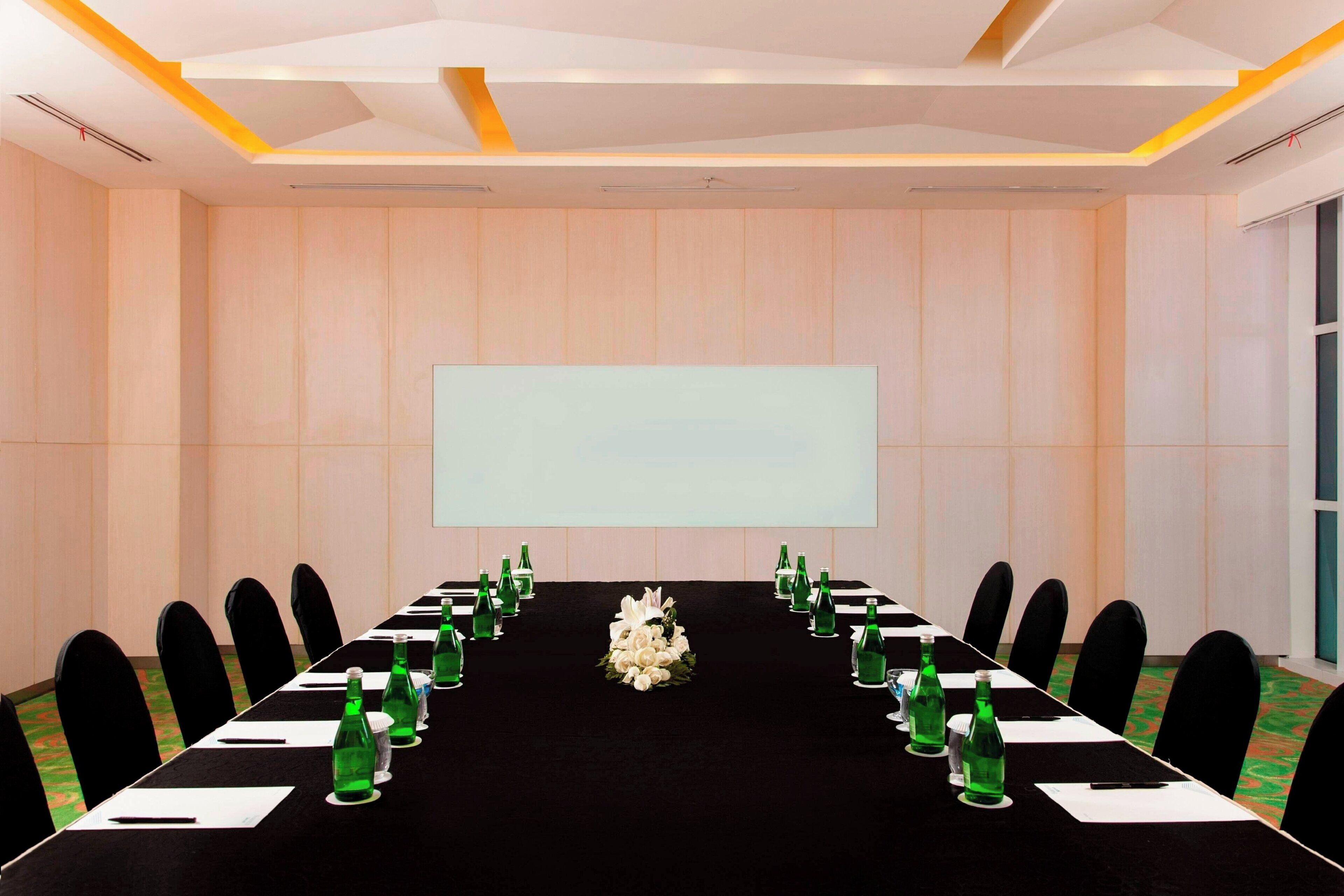 A conference room with a long table, chairs, a projector screen, and a floral centerpiece.