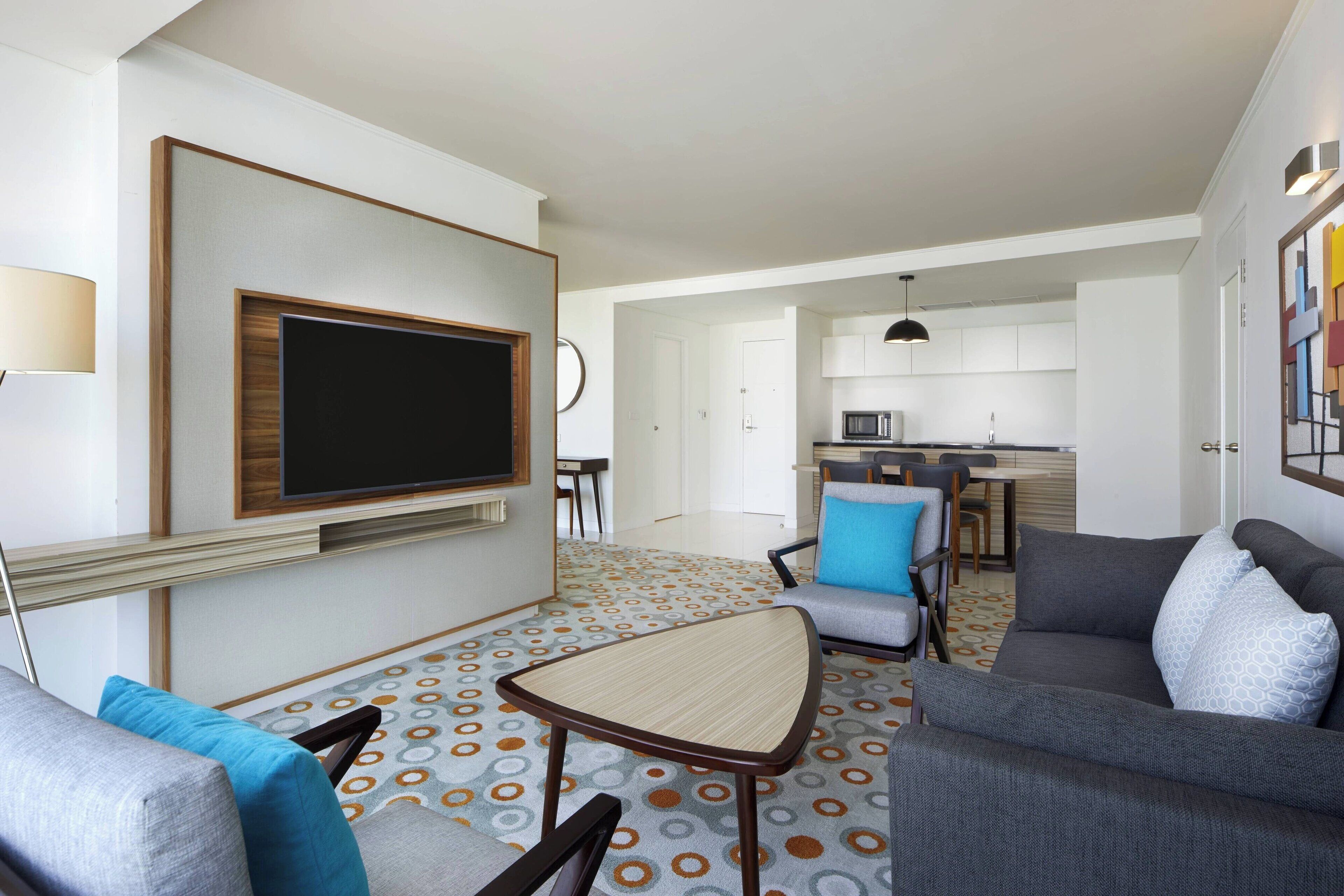 A modern living room with a flat-screen TV, a small coffee table, and a sofa. There is a kitchen area in the background with a microwave and a dining table.