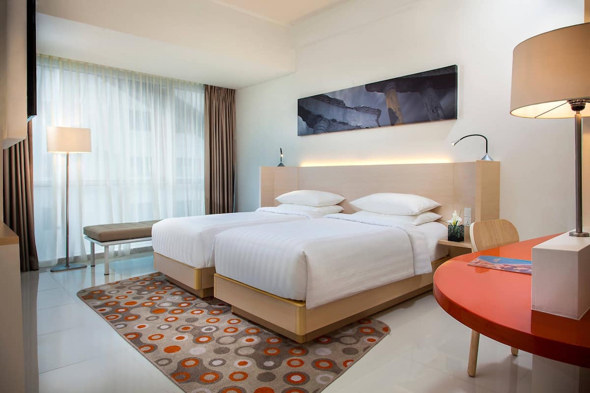 Una habitación de hotel con una cama doble, una mesita de noche con lámpara, una pequeña mesa con una revista y un amplio ventanal con cortinas translúcidas.