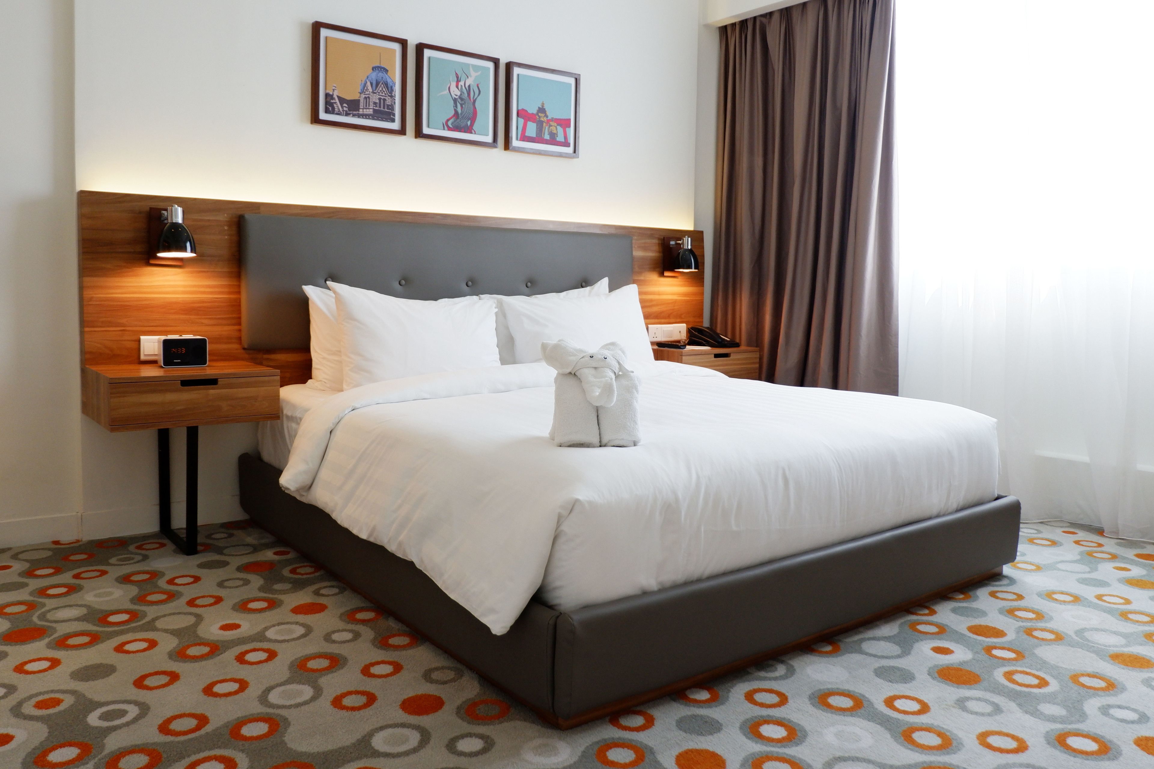 A hotel room with a large bed, two bedside tables, a nightstand, a digital clock, and a wall-mounted lamp.