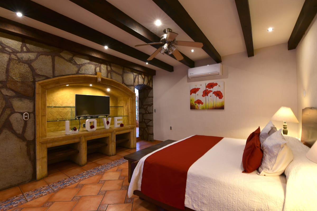 Habitación de hotel con pared de piedra, televisor sobre un soporte de madera, cama con colcha roja y blanca, y un cuadro de flores en la pared.