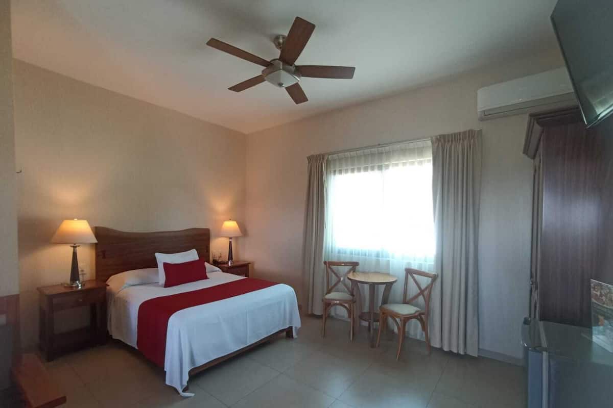 Una habitación de hotel con cama, dos lámparas de noche, ventilador de techo, una pequeña mesa con sillas y televisión.