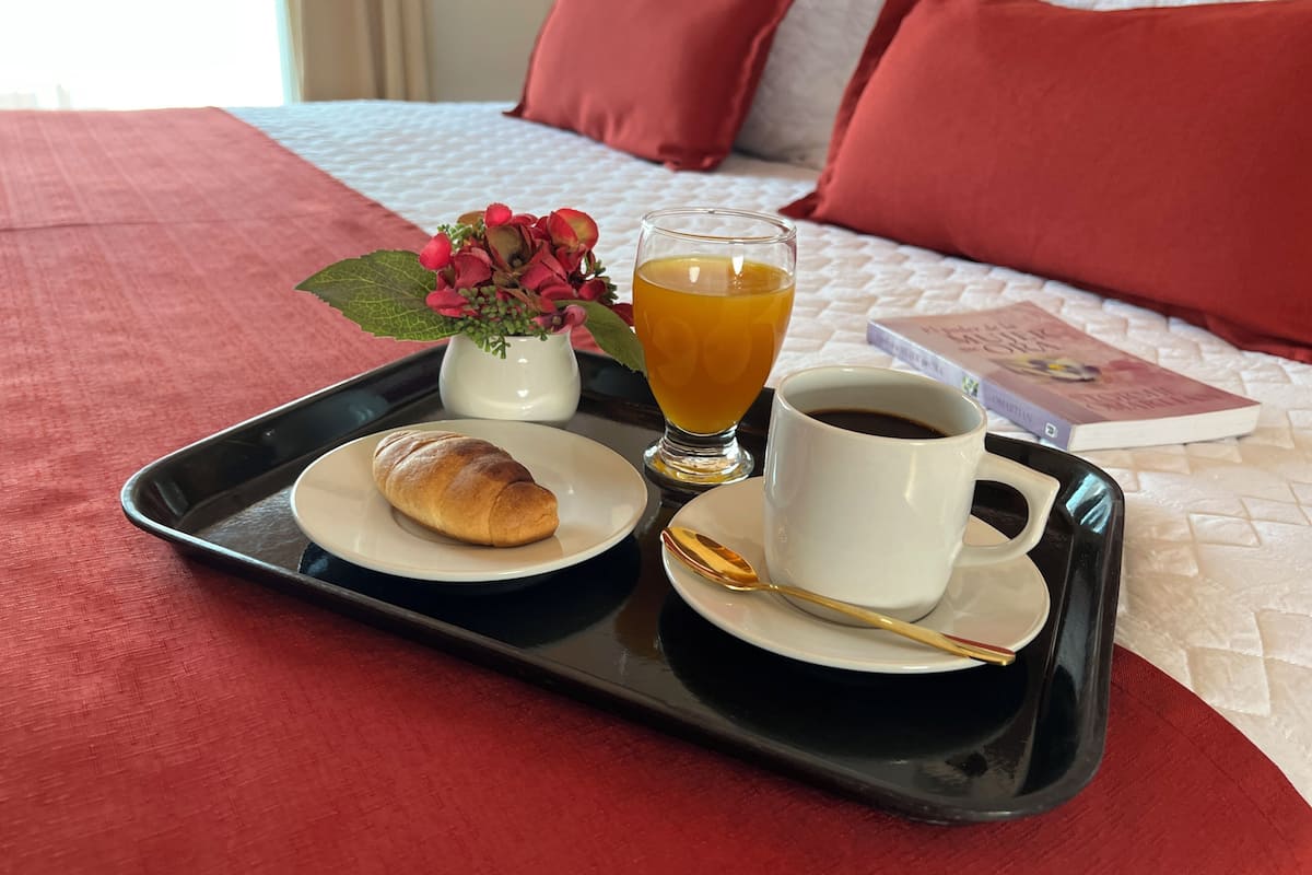 Una bandeja de desayuno con un croissant, un vaso de zumo de naranja y una taza de café sobre una cama con almohadas rojas y una colcha blanca acolchada.