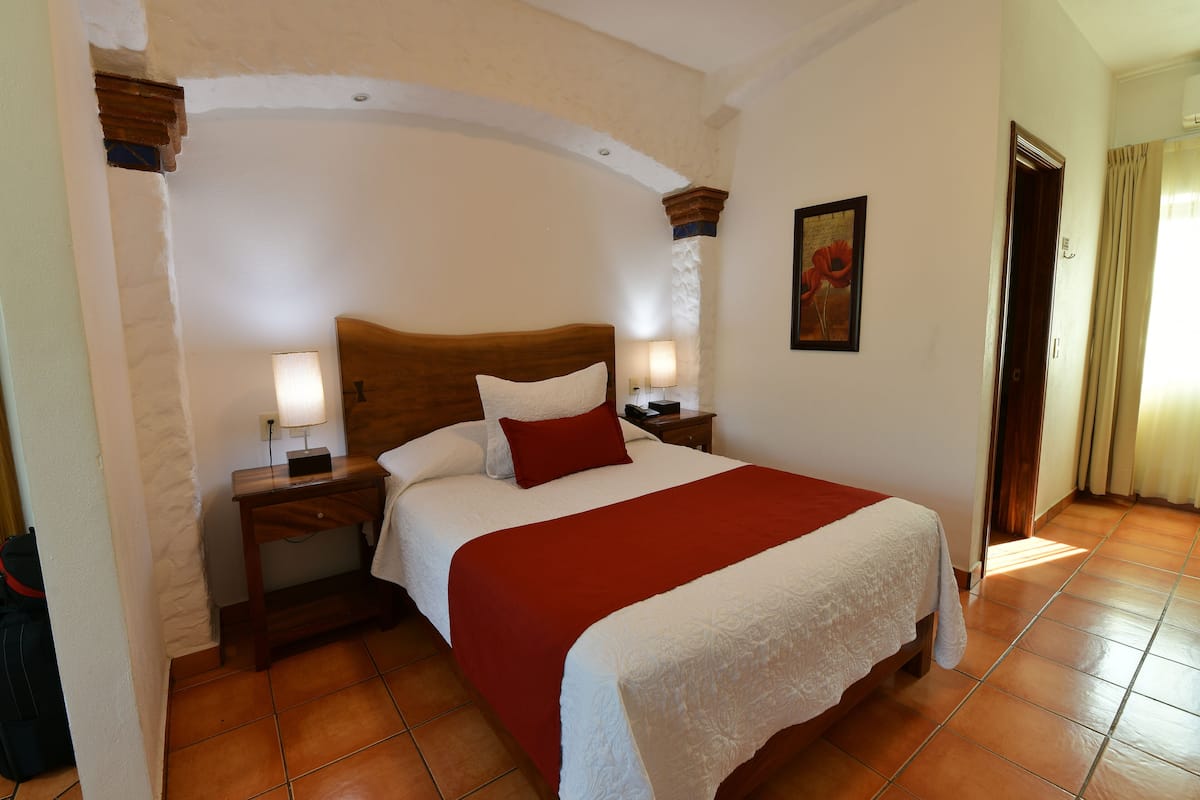 Una habitación de hotel con una cama grande, dos mesitas de noche, una manta roja y un cuadro en la pared.