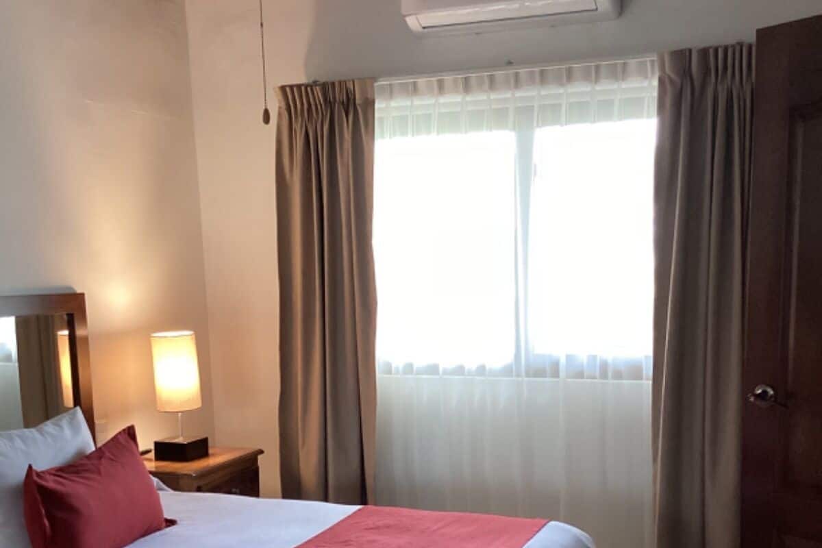Una habitación de hotel con una cama grande, una lámpara de noche, un equipo de aire acondicionado y una ventana con cortinas.