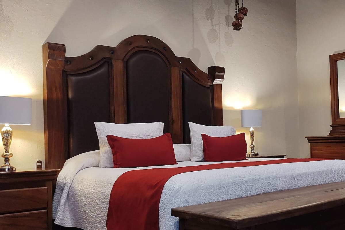 Habitación de hotel ordenada con una cama grande, lámparas de noche, un banco de madera, un aparador y un espejo.